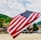 A large American flag is prominently displayed, waving gently against a backdrop of a mountainous landscape. The scene includes several parked cars, wooden buildings, and greenery, suggesting a rural or small-town setting with a clear sky.
