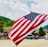 A large American flag is prominently displayed, waving gently against a backdrop of a mountainous landscape. The scene includes several parked cars, wooden buildings, and greenery, suggesting a rural or small-town setting with a clear sky.
