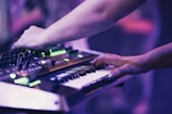 Hands are interacting with a synthesizer and keyboard, creating an immersive musical experience. The image is bathed in soft purple and green lighting, emphasizing the electronic equipment.