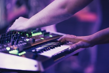 Hands are interacting with a synthesizer and keyboard, creating an immersive musical experience. The image is bathed in soft purple and green lighting, emphasizing the electronic equipment.