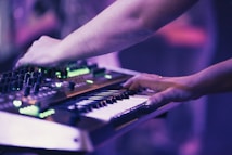 Hands are interacting with a synthesizer and keyboard, creating an immersive musical experience. The image is bathed in soft purple and green lighting, emphasizing the electronic equipment.