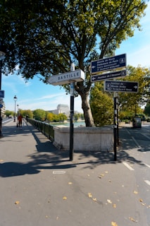 Several street signs are situated on a paved pathway surrounded by trees and greenery. The main sign prominently displays the word 'Bastille' while other directional signs point towards different Parisian locations such as 'Mairie de Paris' and 'Institut du Monde Arabe'. A person is walking along the sidewalk, and a clear blue sky is visible above.