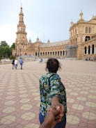 A person with a floral-patterned shirt is leading the viewer by the hand across a wide, tiled plaza with a grand, historic building featuring arches and a tower in the background. Several other people are visible walking in the distance.