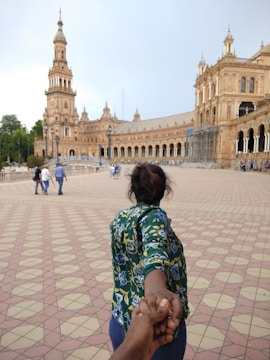 A person with a floral-patterned shirt is leading the viewer by the hand across a wide, tiled plaza with a grand, historic building featuring arches and a tower in the background. Several other people are visible walking in the distance.