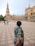 A person with a floral-patterned shirt is leading the viewer by the hand across a wide, tiled plaza with a grand, historic building featuring arches and a tower in the background. Several other people are visible walking in the distance.