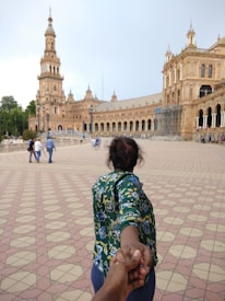 A person with a floral-patterned shirt is leading the viewer by the hand across a wide, tiled plaza with a grand, historic building featuring arches and a tower in the background. Several other people are visible walking in the distance.