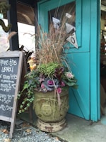 A large, ornate planter is filled with vibrant plants, including ornamental cabbages and tall grasses, placed next to a teal-colored wooden door. A blackboard with white handwriting displays prices for mums. The setting appears to be outside a shop or greenhouse.