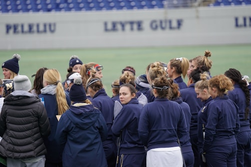 A group of people, mostly women, stand together on a sports field. They are wearing dark blue jackets and some have ponytails or buns. The background features a partially visible sign with the words 'PELELIU' and 'LEYTE GULF' on a wall.