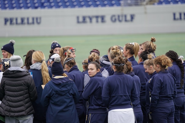 A group of people, mostly women, stand together on a sports field. They are wearing dark blue jackets and some have ponytails or buns. The background features a partially visible sign with the words 'PELELIU' and 'LEYTE GULF' on a wall.