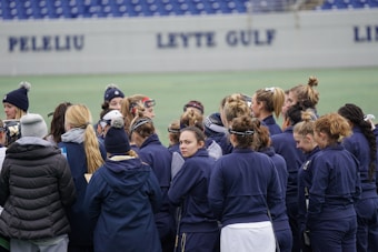 A group of people, mostly women, stand together on a sports field. They are wearing dark blue jackets and some have ponytails or buns. The background features a partially visible sign with the words 'PELELIU' and 'LEYTE GULF' on a wall.
