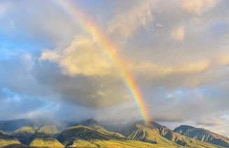 A vibrant rainbow arching over green mountain slopes under a cloudy sky