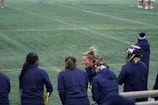 A group of athletes wearing navy jackets stands on a sports field. One person in the center appears to be laughing joyfully, while others are engaged in conversation or looking elsewhere. A player with a helmet is visible on the right, and another group is seen in the background on the field. The field is covered in green artificial turf with yellow and blue lines marking boundaries.
