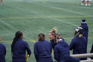 A group of athletes wearing navy jackets stands on a sports field. One person in the center appears to be laughing joyfully, while others are engaged in conversation or looking elsewhere. A player with a helmet is visible on the right, and another group is seen in the background on the field. The field is covered in green artificial turf with yellow and blue lines marking boundaries.