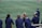 A group of athletes wearing navy jackets stands on a sports field. One person in the center appears to be laughing joyfully, while others are engaged in conversation or looking elsewhere. A player with a helmet is visible on the right, and another group is seen in the background on the field. The field is covered in green artificial turf with yellow and blue lines marking boundaries.
