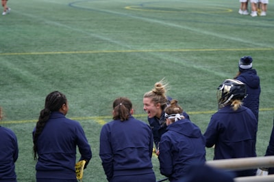 A group of athletes wearing navy jackets stands on a sports field. One person in the center appears to be laughing joyfully, while others are engaged in conversation or looking elsewhere. A player with a helmet is visible on the right, and another group is seen in the background on the field. The field is covered in green artificial turf with yellow and blue lines marking boundaries.
