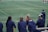 A group of athletes wearing navy jackets stands on a sports field. One person in the center appears to be laughing joyfully, while others are engaged in conversation or looking elsewhere. A player with a helmet is visible on the right, and another group is seen in the background on the field. The field is covered in green artificial turf with yellow and blue lines marking boundaries.