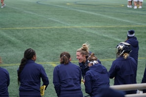 A group of athletes wearing navy jackets stands on a sports field. One person in the center appears to be laughing joyfully, while others are engaged in conversation or looking elsewhere. A player with a helmet is visible on the right, and another group is seen in the background on the field. The field is covered in green artificial turf with yellow and blue lines marking boundaries.
