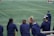 A group of athletes wearing navy jackets stands on a sports field. One person in the center appears to be laughing joyfully, while others are engaged in conversation or looking elsewhere. A player with a helmet is visible on the right, and another group is seen in the background on the field. The field is covered in green artificial turf with yellow and blue lines marking boundaries.