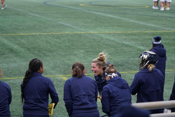 A group of athletes wearing navy jackets stands on a sports field. One person in the center appears to be laughing joyfully, while others are engaged in conversation or looking elsewhere. A player with a helmet is visible on the right, and another group is seen in the background on the field. The field is covered in green artificial turf with yellow and blue lines marking boundaries.