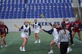 A women's lacrosse game is taking place on a large stadium field. Several players in maroon jerseys marked with 'Saint Joseph's' face off against players in white jerseys labeled 'Navy'. The athletes are actively engaged, with one player in 'Navy' about to catch or throw the ball. Another player attempts to block using her stick. A referee in a black and white striped shirt is visible in the foreground. The backdrop includes empty blue stadium seats.