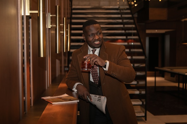 A well-dressed man stands in an elegantly designed interior, holding a drink in one hand while resting his other hand on a newspaper placed on the counter. The setting includes modern lighting fixtures and a staircase leading upwards in the background. The atmosphere exudes a sense of sophistication and comfort.