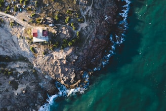 An aerial view of a rugged coastline where a solitary house with a red roof is nestled among rocky terrain. The rocky cliffs meet the deep blue ocean, with waves crashing against the shore. Sparse vegetation is visible, enhancing the natural and remote feel of the location.