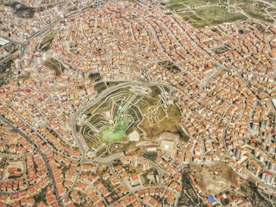 An aerial view of a densely populated urban area with numerous buildings featuring terracotta roofs. In the center, a large green space with a park or garden is visible, featuring pathways and patches of greenery. Roads and streets weave throughout the city, connecting different neighborhoods.