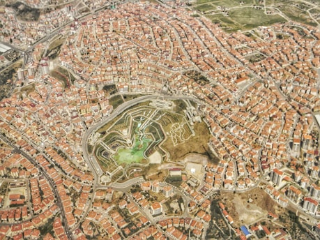 An aerial view of a densely populated urban area with numerous buildings featuring terracotta roofs. In the center, a large green space with a park or garden is visible, featuring pathways and patches of greenery. Roads and streets weave throughout the city, connecting different neighborhoods.