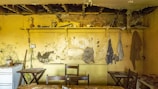 An old, dilapidated kitchen with peeling paint and deteriorating walls. A shelf holds various objects such as vases and a candle holder. The ceiling is damaged and partially exposes the beams. Chairs and tables are scattered around, and a small stove with a kettle sits in the corner. There are hooks on the wall with hanging cloths or aprons.