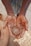 Hands covered in clay hold a small, newly formed clay pot, suggesting a scene of pottery making. The texture of the wet clay is prominent, and the background hints at more pottery items.