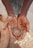Hands covered in clay hold a small, newly formed clay pot, suggesting a scene of pottery making. The texture of the wet clay is prominent, and the background hints at more pottery items.
