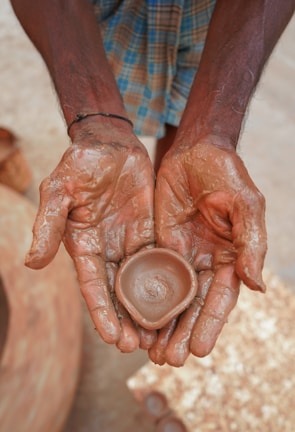 Hands covered in clay hold a small, newly formed clay pot, suggesting a scene of pottery making. The texture of the wet clay is prominent, and the background hints at more pottery items.