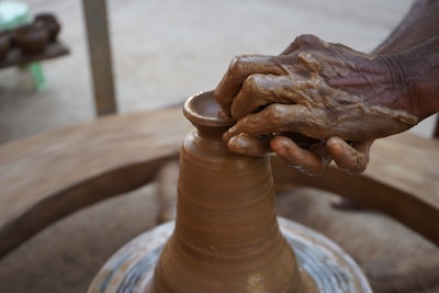 A skilled pair of hands is shaping a clay pot on a spinning pottery wheel. The hands are covered in wet clay, suggesting a close engagement with the material. In the background, there are blurred items, possibly indicating an outdoor or workshop setting.