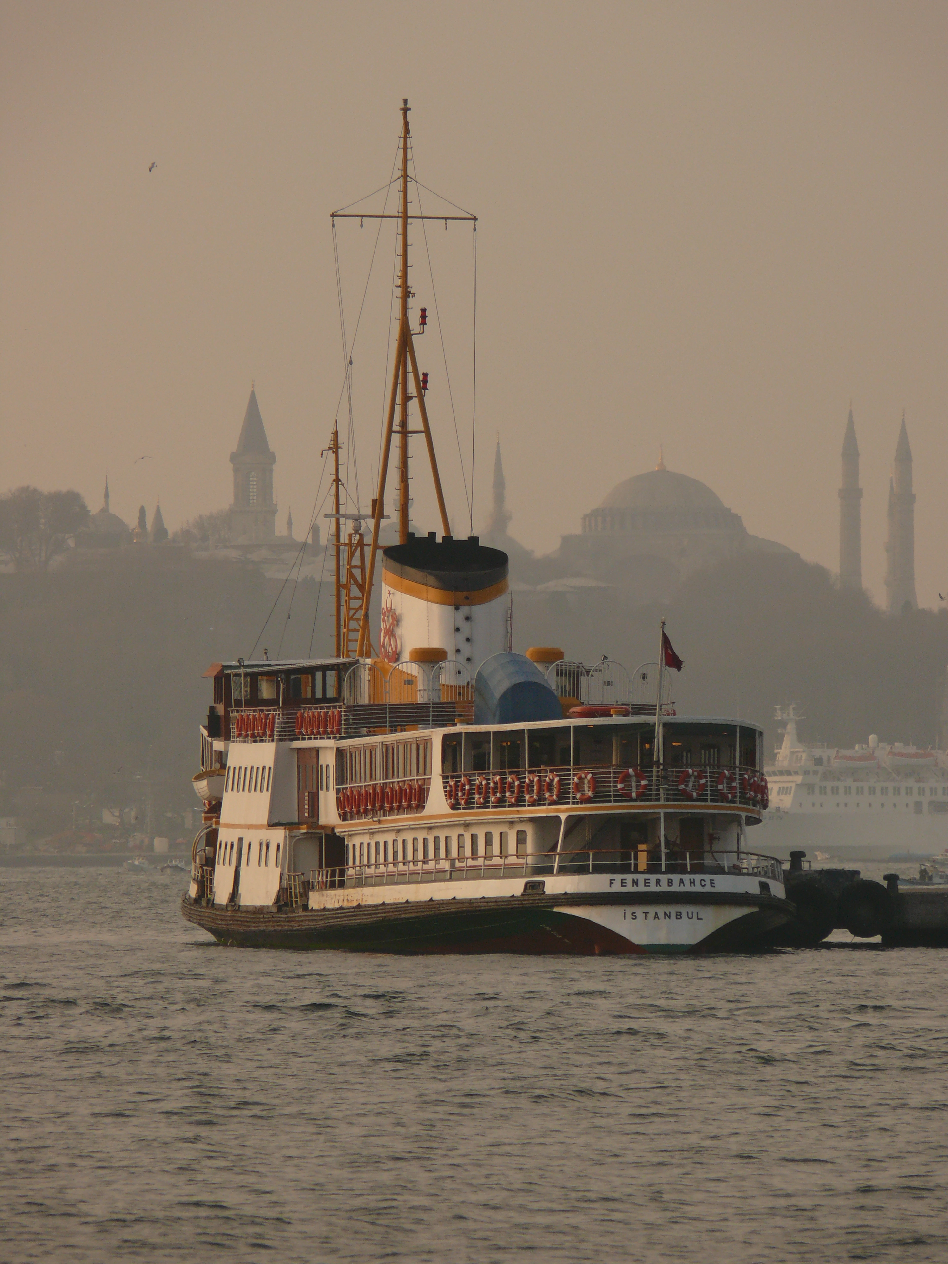 A classic white-and-black Istanbul city ferry on the water in soft pink haze, the domes and minarets of the historic peninsula rising faintly behind it