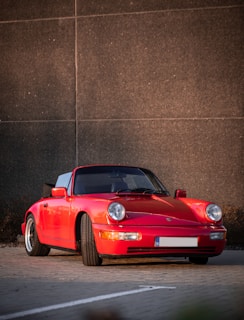 A classic red sports car with a sleek, aerodynamic design is parked in front of a textured, dark concrete wall. The car's polished surface reflects sunlight, highlighting its curves and contours. It sits on wide tires with intricate rims and its headlights and turn signals are clearly visible.