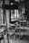 A monochrome photograph of a cafe interior featuring wooden tables and chairs. The cafe has large windows allowing natural light to stream in, and there are hanging plants visible near the window. The counter area in the background is partially visible, with various items and a menu board hanging above.