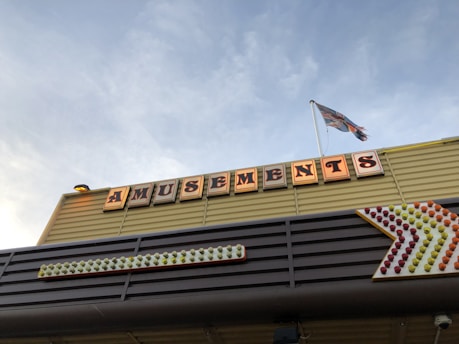 A structure with a sign displaying the word 'Amusements' illuminated in large, vintage-style letters. Beneath the sign is an arrow made of multicolored lights pointing to the right. A flag flutters on a pole above the sign against a backdrop of a clear blue sky.