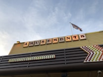 A structure with a sign displaying the word 'Amusements' illuminated in large, vintage-style letters. Beneath the sign is an arrow made of multicolored lights pointing to the right. A flag flutters on a pole above the sign against a backdrop of a clear blue sky.