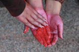 Hand holding a bowl of pure red henna powder called 'مسحوق الحناء النقي' against a warm Moroccan textile