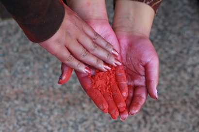 A close-up of a pair of hands holding a vivid red powder. The hands are slightly stained with the red pigment, suggesting involvement in an activity related to this substance. The background is a blurred gray and black surface.