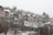 Snow is falling heavily on a hillside village capturing several houses with sloped roofs. The roofs and ground are dusted with snow, and bare trees are visible in the foreground, adding a wintry feel to the scene.