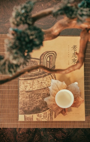 A cup of tea sits on a textured paper with Asian-inspired artwork and text, positioned on a bamboo mat. Above, blurred branches or twigs create a natural frame.
