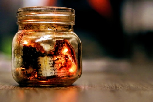 A glass jar on a wooden surface contains burning paper, creating a warm glow. The background is blurred, emphasizing the flaming papers inside the jar.