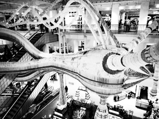 A high-contrast black and white image of a large, ornate indoor structure resembling an abstract sculpture or chandelier. The setting appears to be inside a multi-level retail store or mall with escalators and various lighting elements. People are visible in the background, engaged in shopping and looking around.