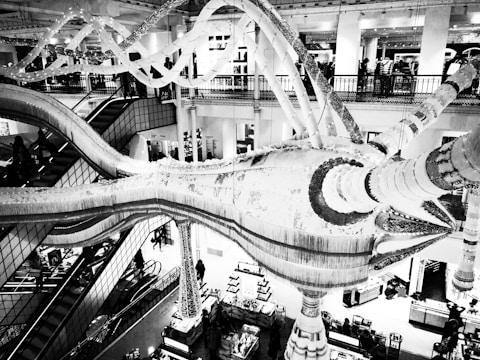 A high-contrast black and white image of a large, ornate indoor structure resembling an abstract sculpture or chandelier. The setting appears to be inside a multi-level retail store or mall with escalators and various lighting elements. People are visible in the background, engaged in shopping and looking around.