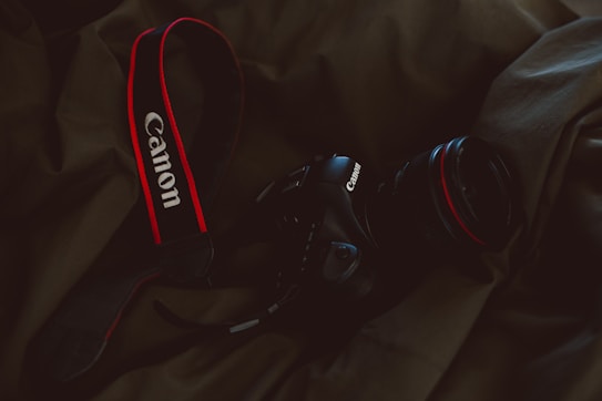 A Canon camera with a red and black strap placed on a dark fabric background. The camera lens is facing sideways and the brand name is visible on both the strap and the camera body.