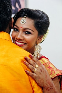 A woman wearing traditional attire is smiling warmly as she embraces someone. She has intricate jewelry, including earrings and a prominent headpiece. Her hand, adorned with henna and jewelry, is visible on the person's shoulder.
