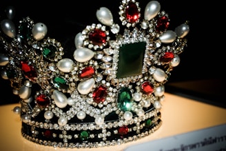 A luxurious crown adorned with an array of precious gemstones and pearls. The design features large red and green stones surrounded by a multitude of smaller clear crystals, creating an intricate and opulent pattern.