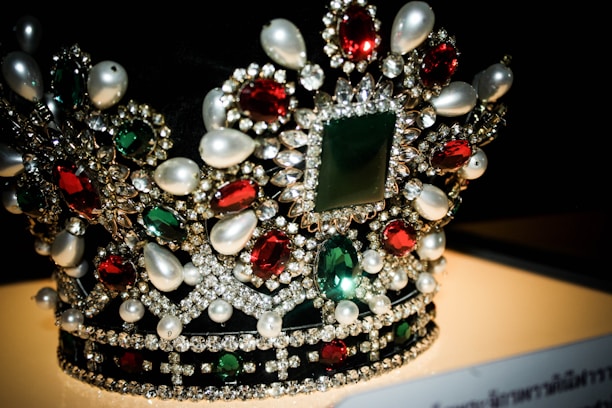 A luxurious crown adorned with an array of precious gemstones and pearls. The design features large red and green stones surrounded by a multitude of smaller clear crystals, creating an intricate and opulent pattern.