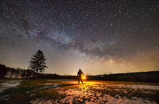 A solitary figure cloaked in gray, holding a glowing lantern, standing atop a snowy mountain under a starry night.